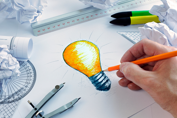 A hand drawn picture of a bright light bulb surrounded with crumpled pieces of paper, pens, ruler and a compass.